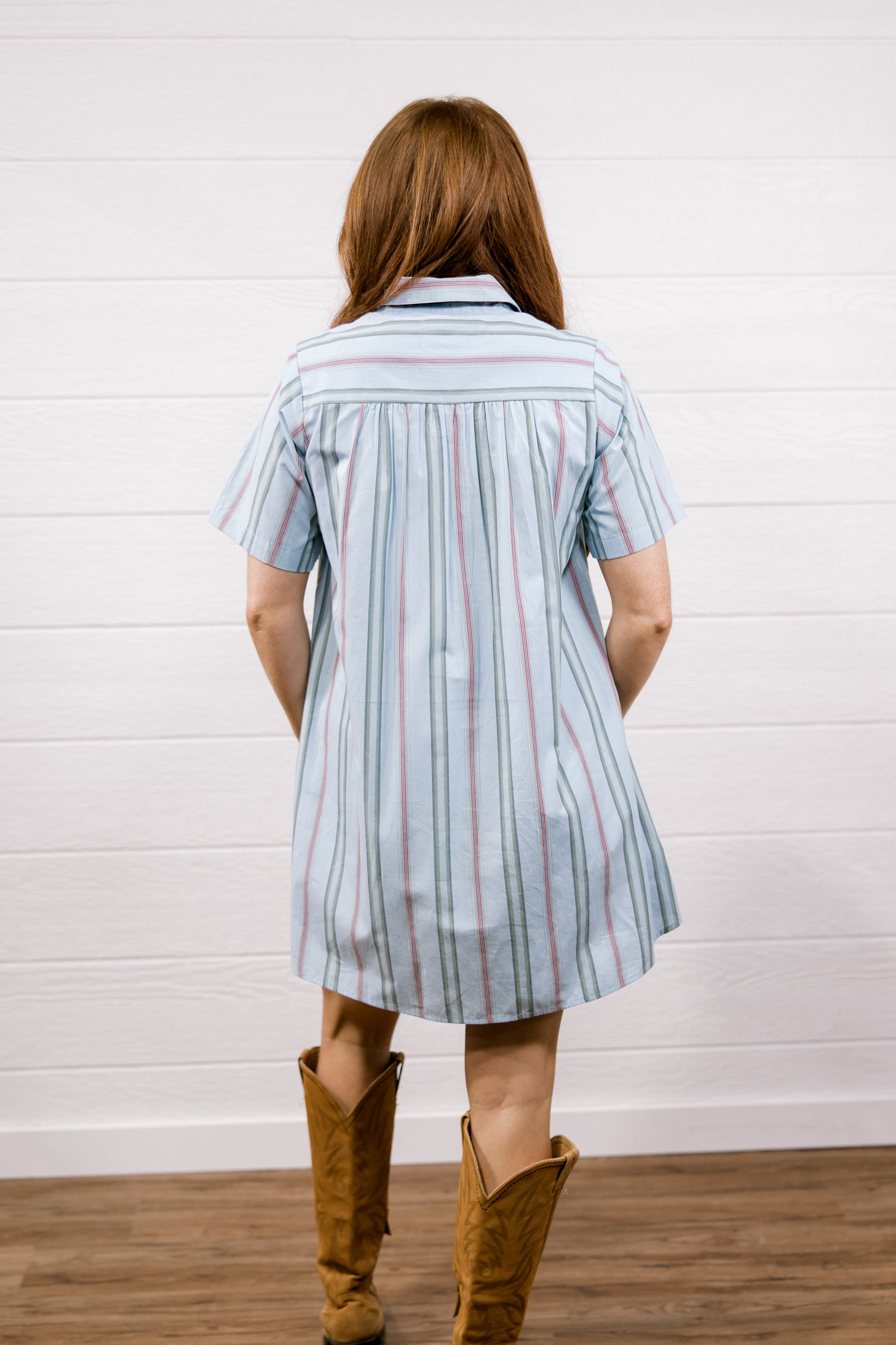 A woman viewed from behind wearing a light blue short-sleeve striped shirt dress with thin red and white pinstripes, paired with brown cowboy boots, standing against a white paneled wall on a wood floor.