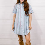 A woman modeling a light blue short-sleeve button-up shirt dress with vertical red and white stripes, paired with tan cowboy boots, standing against a white wood-paneled wall on a wooden floor.