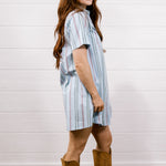 A woman modeling a short-sleeve light blue striped shirt dress with brown cowboy boots, standing against a white wood-paneled wall on a wooden floor.