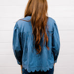 A woman with long, wavy light brown hair is shown from behind, wearing a blue and white vertically striped long-sleeve blouse with a scalloped hem and dark blue jeans, standing against a white wood-paneled wall.