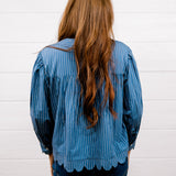 A woman with long, wavy light brown hair is shown from behind, wearing a blue and white vertically striped long-sleeve blouse with a scalloped hem and dark blue jeans, standing against a white wood-paneled wall.