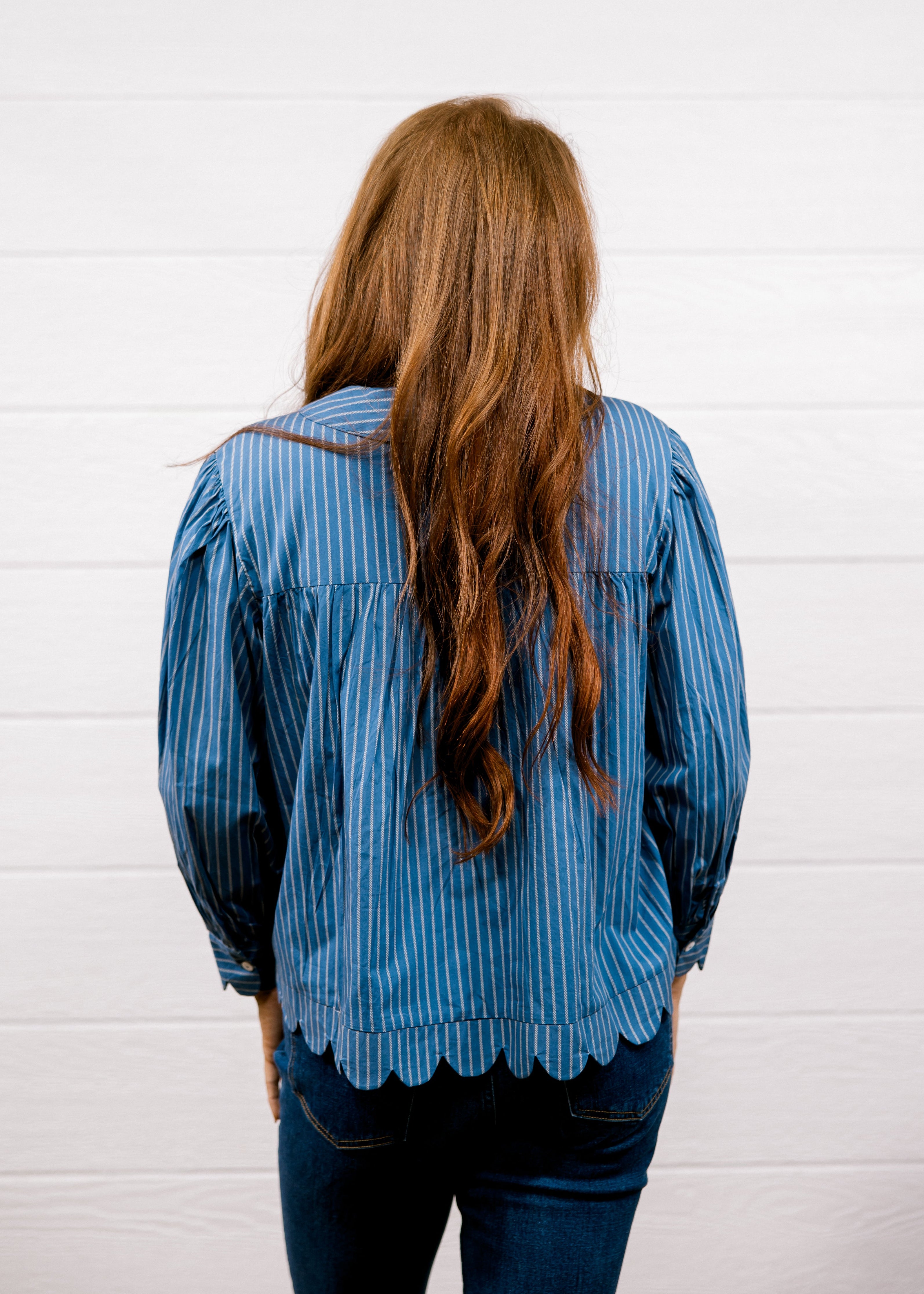 A woman with long, wavy light brown hair is shown from behind, wearing a blue and white vertically striped long-sleeve blouse with a scalloped hem and dark blue jeans, standing against a white wood-paneled wall.