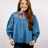 A smiling young woman with long brown hair stands against a white paneled wall wearing a blue vertically striped blouse with colorful embroidered detailing on the chest and dark blue jeans.
