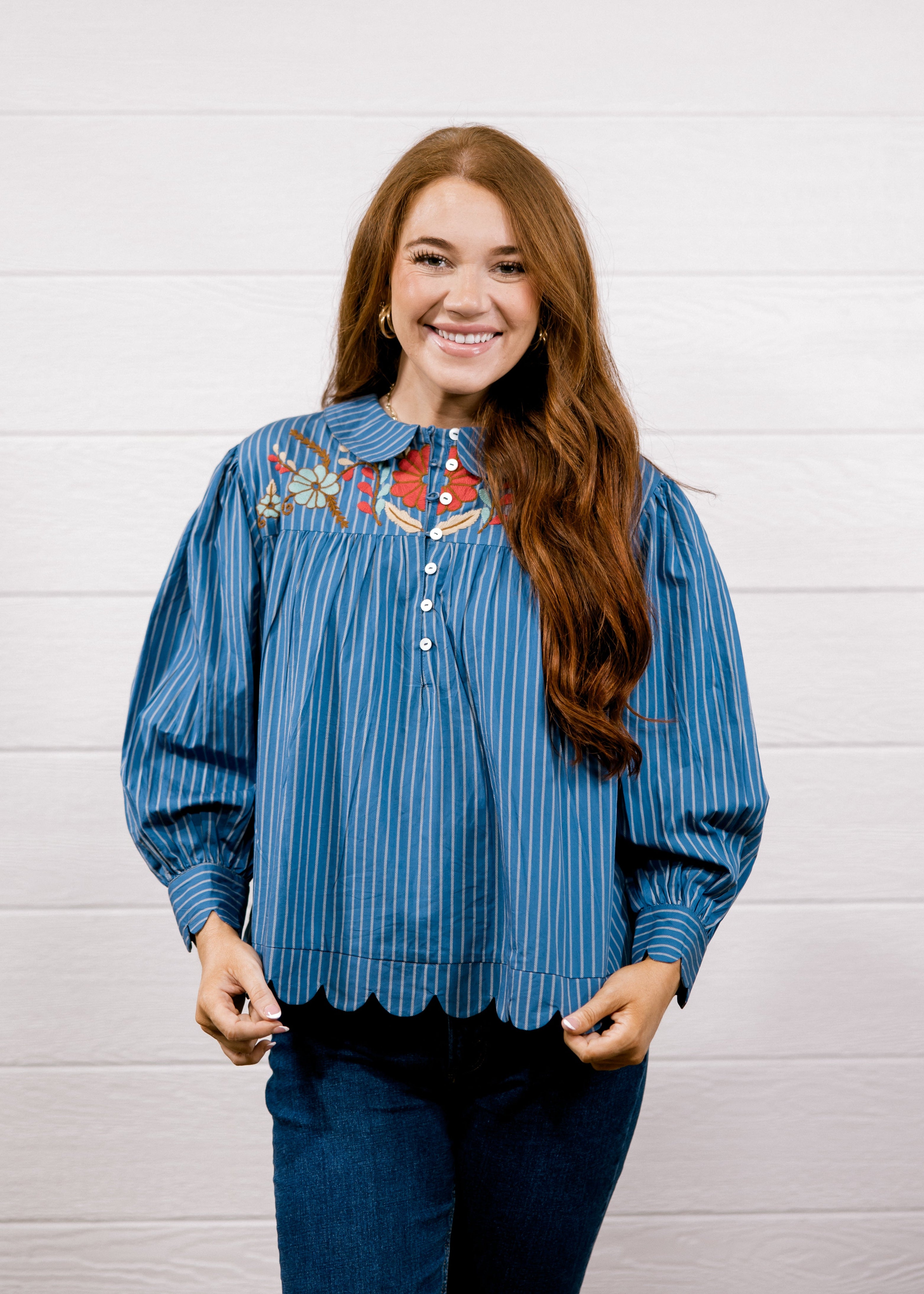 A smiling young woman with long brown hair stands against a white paneled wall wearing a blue vertically striped blouse with colorful embroidered detailing on the chest and dark blue jeans.