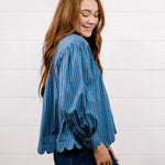 A woman with long red hair is shown from the side, wearing a blue and navy vertically striped long-sleeve blouse with scalloped edges and dark blue jeans, standing against a white wood-paneled wall.