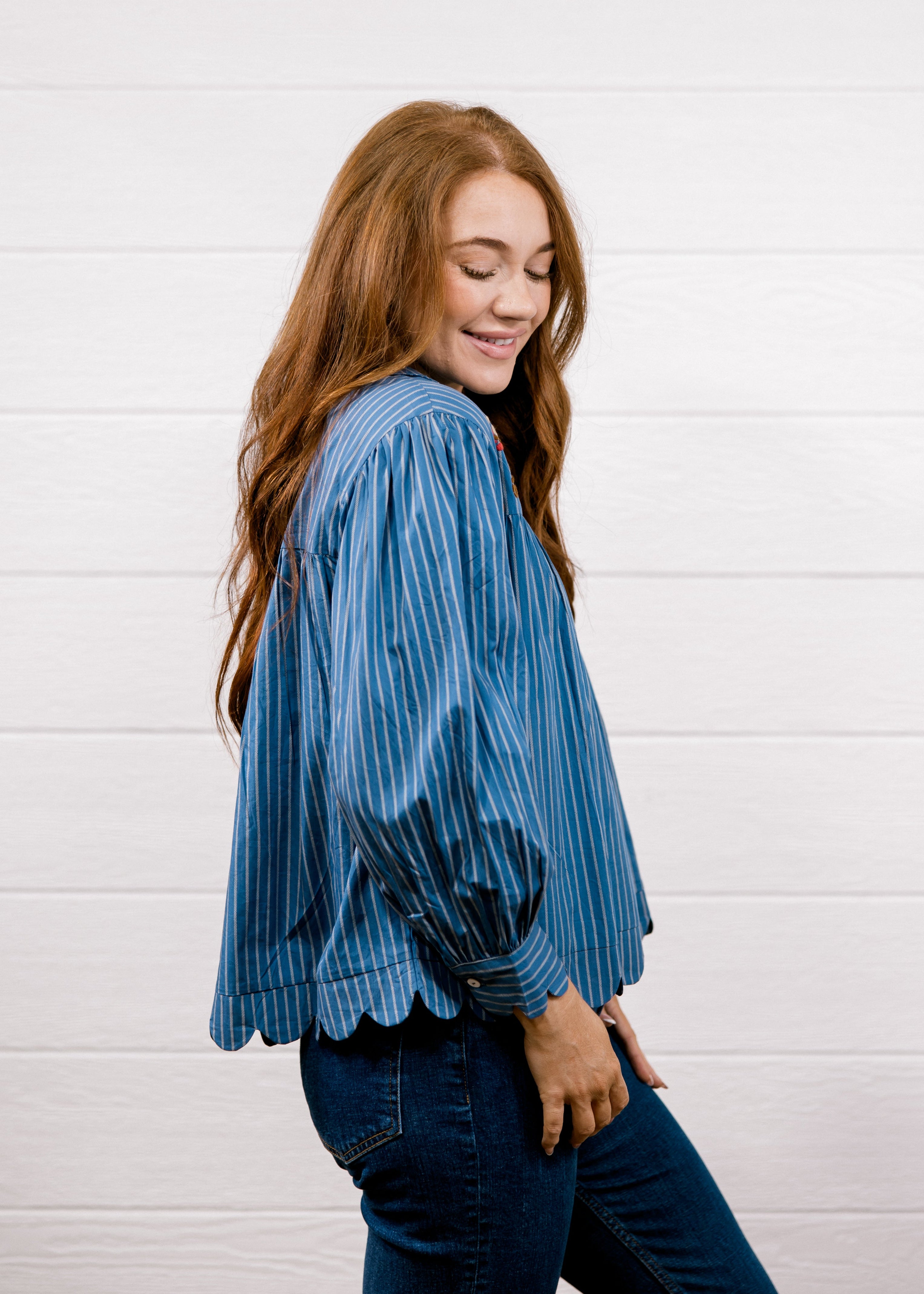 A woman with long red hair is shown from the side, wearing a blue and navy vertically striped long-sleeve blouse with scalloped edges and dark blue jeans, standing against a white wood-paneled wall.