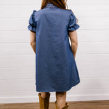 A woman viewed from behind wearing a blue short-sleeve linen shirt dress with ruffled shoulders and tan cowboy boots, standing on a wooden floor against a white shiplap wall.