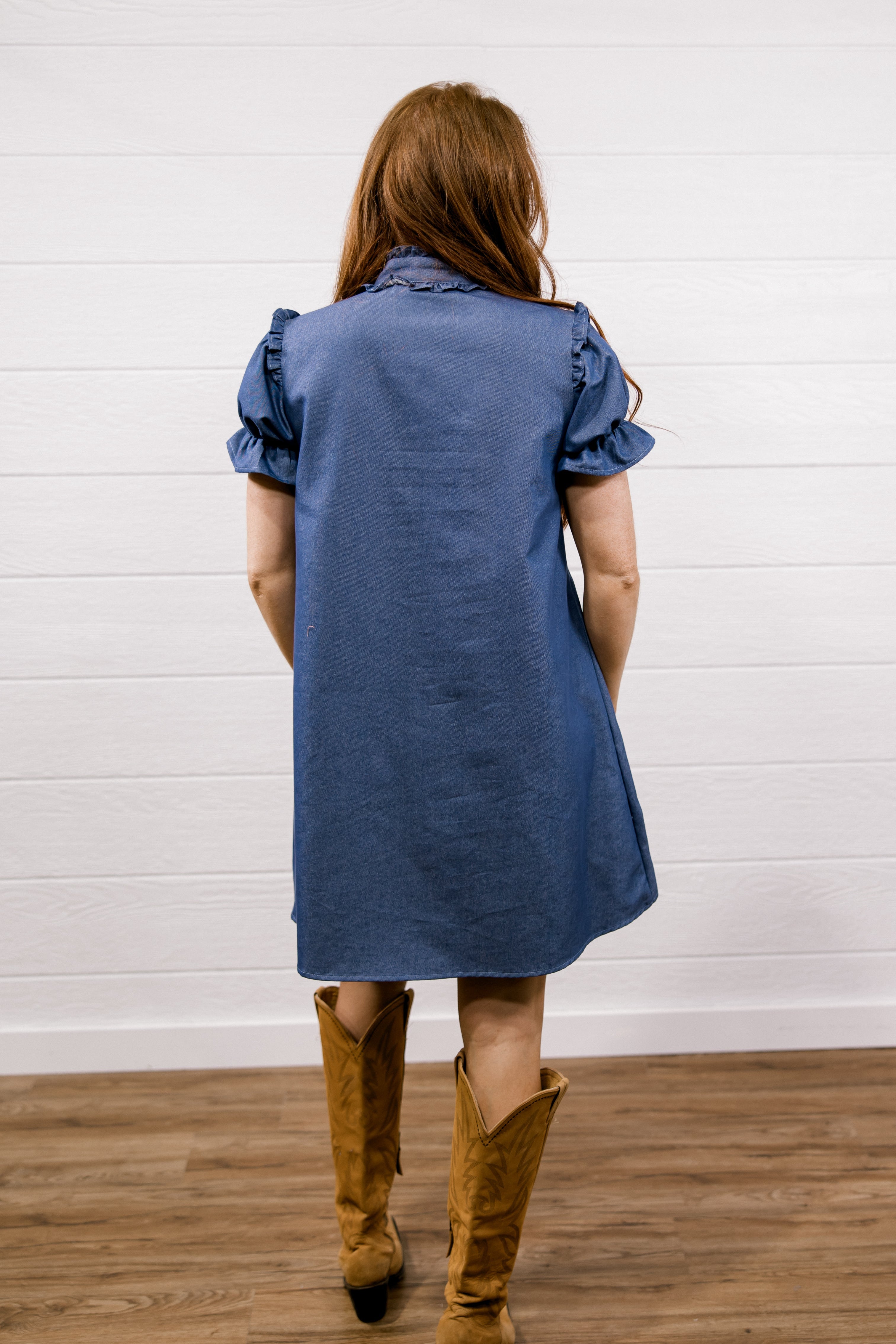 A woman viewed from behind wearing a blue short-sleeve linen shirt dress with ruffled shoulders and tan cowboy boots, standing on a wooden floor against a white shiplap wall.