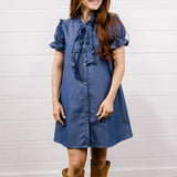A woman wearing a blue denim-style short-sleeve dress with a ruffled tie neckline and brown suede cowboy boots, standing against a white shiplap wall and wood floor.