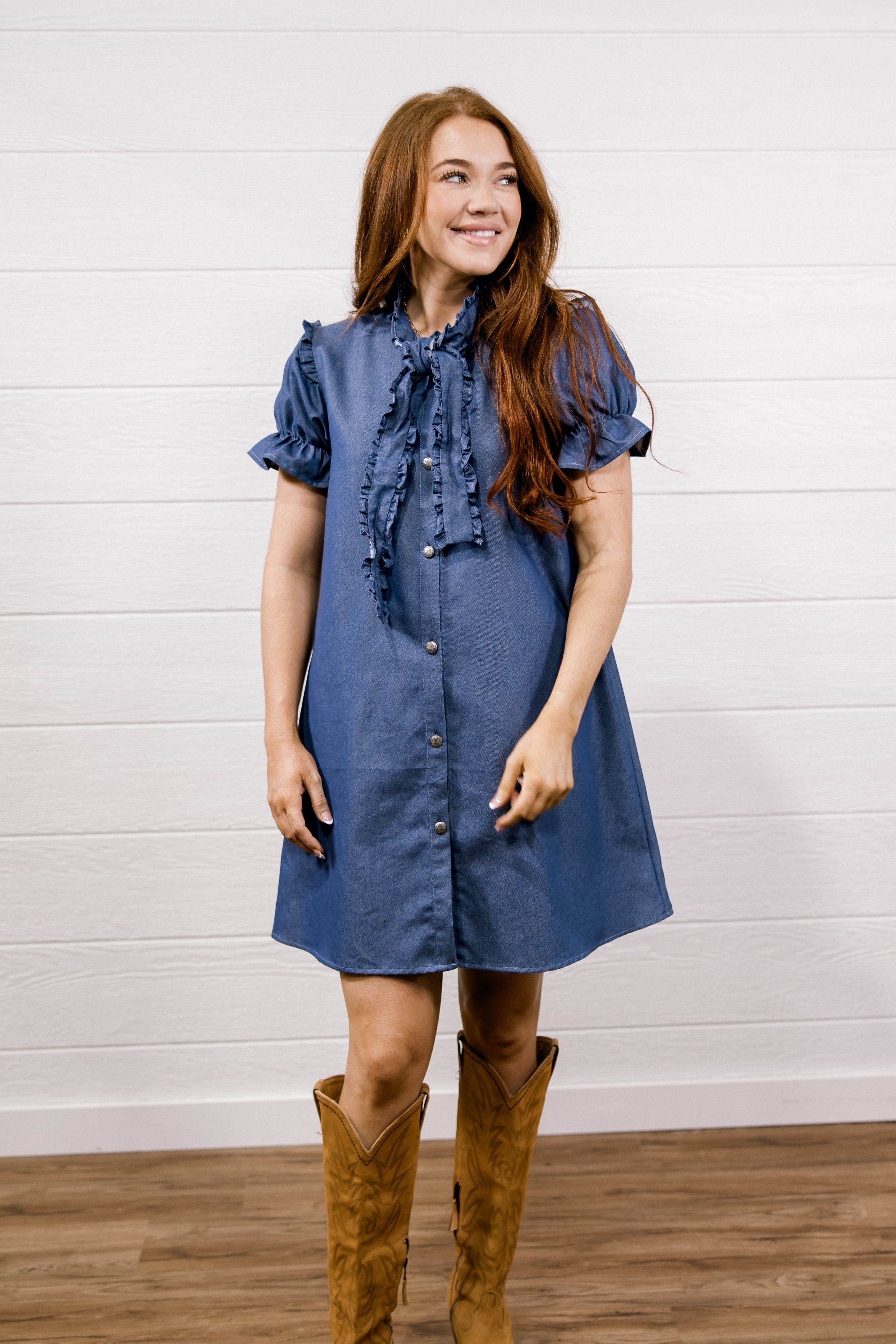 A woman wearing a blue denim-style short-sleeve dress with a ruffled tie neckline and brown suede cowboy boots, standing against a white shiplap wall and wood floor.