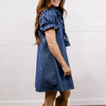 A woman with long red hair stands in profile wearing a blue short-sleeve dress with ruffled trim and tan cowboy boots, posed against a white shiplap wall and wood floor.