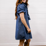 A woman with long red hair stands in profile wearing a blue short-sleeve dress with ruffled trim and tan cowboy boots, posed against a white shiplap wall and wood floor.