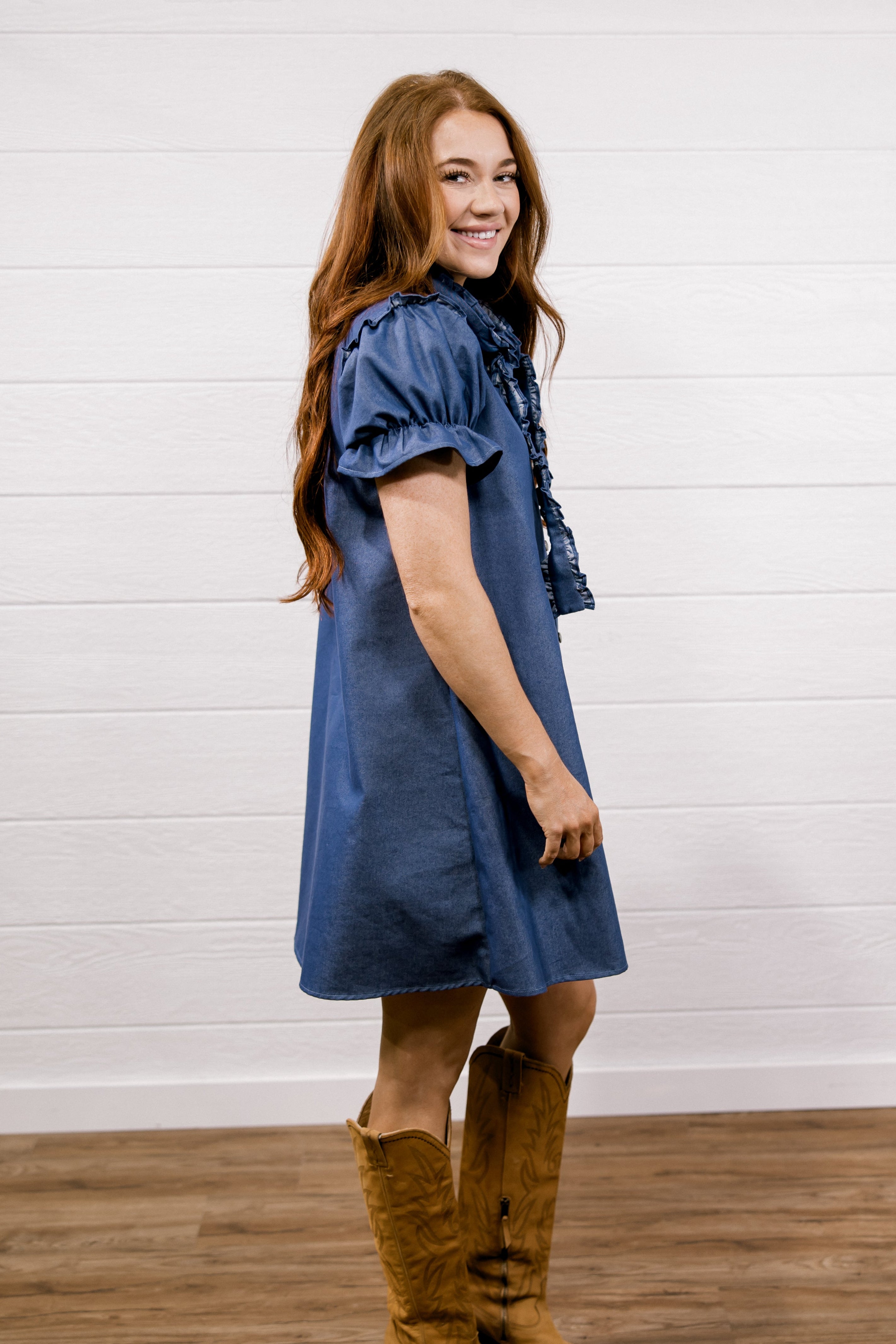A woman with long red hair stands in profile wearing a blue short-sleeve dress with ruffled trim and tan cowboy boots, posed against a white shiplap wall and wood floor.