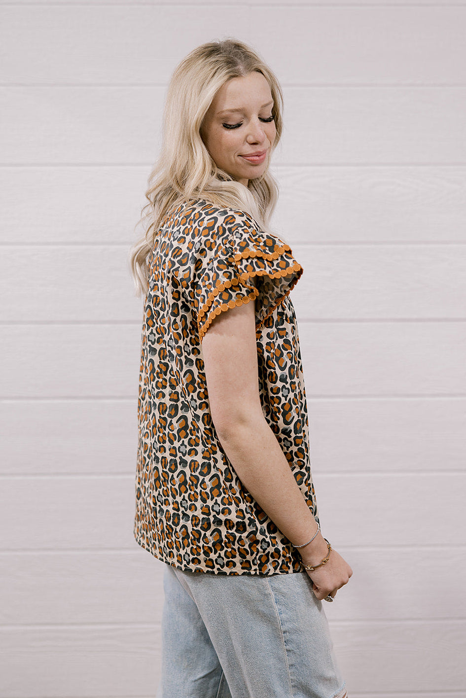 A woman wearing a leopard print short-sleeve top with ruffled sleeves and light-wash denim shorts, standing against a white wood-paneled wall.