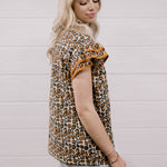 A woman wearing a leopard print short-sleeve top with ruffled sleeves and light-wash denim shorts, standing against a white wood-paneled wall.