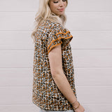 A woman wearing a leopard print short-sleeve top with ruffled sleeves and light-wash denim shorts, standing against a white wood-paneled wall.