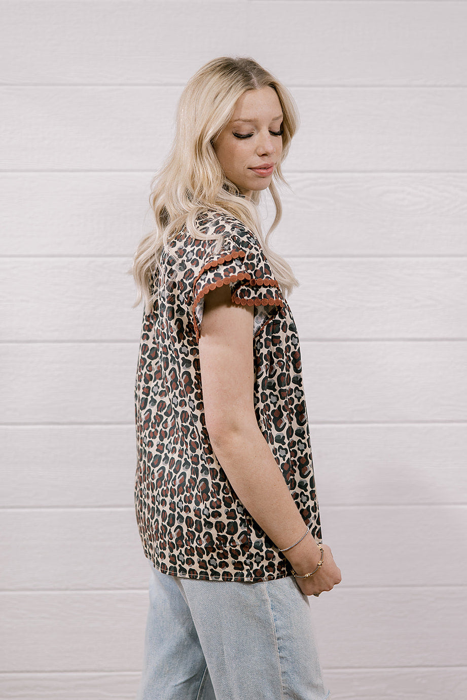 A woman with blonde hair is shown from the side, wearing a brown and beige leopard-print short-sleeve top with brown ruffle trim at the cuffs, paired with light-wash distressed jeans, set against a white wood-paneled wall.