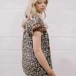 A woman with blonde hair is shown from the side, wearing a brown and beige leopard-print short-sleeve top with brown ruffle trim at the cuffs, paired with light-wash distressed jeans, set against a white wood-paneled wall.