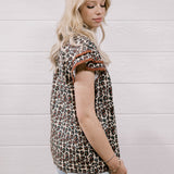 A woman with blonde hair is shown from the side, wearing a brown and beige leopard-print short-sleeve top with brown ruffle trim at the cuffs, paired with light-wash distressed jeans, set against a white wood-paneled wall.