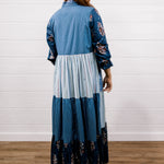 A woman viewed from the back wearing a long patchwork maxi dress with blue and white stripes, solid light blue panels, and floral print sleeves and hem, standing on a wood floor against a white shiplap wall.
