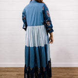 A woman viewed from the back wearing a long patchwork maxi dress with blue and white stripes, solid light blue panels, and floral print sleeves and hem, standing on a wood floor against a white shiplap wall.