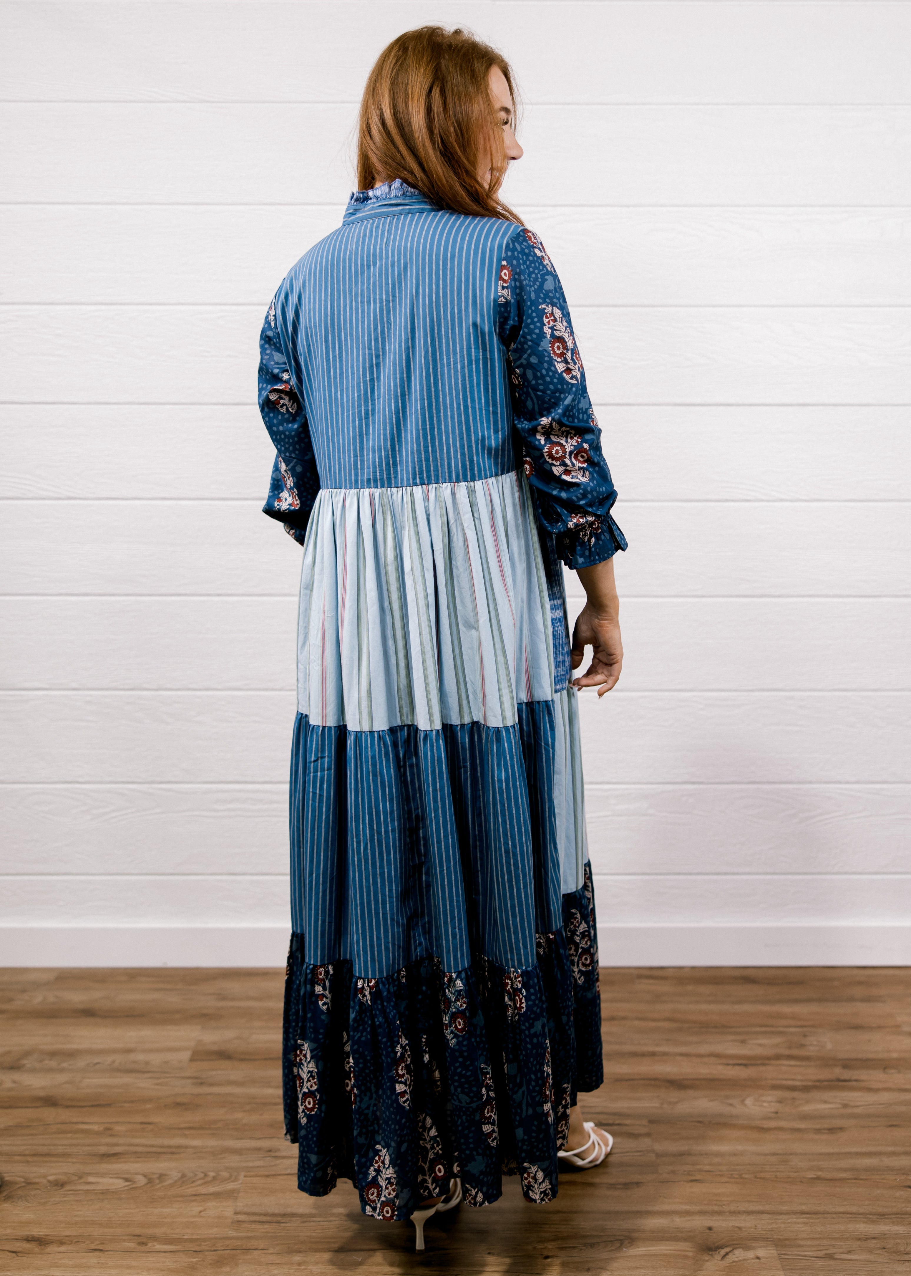 A woman viewed from the back wearing a long patchwork maxi dress with blue and white stripes, solid light blue panels, and floral print sleeves and hem, standing on a wood floor against a white shiplap wall.