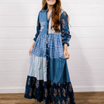 A woman modeling a long-sleeve patchwork maxi dress with blue plaid, striped, and floral prints, standing against a white shiplap wall on a wooden floor.