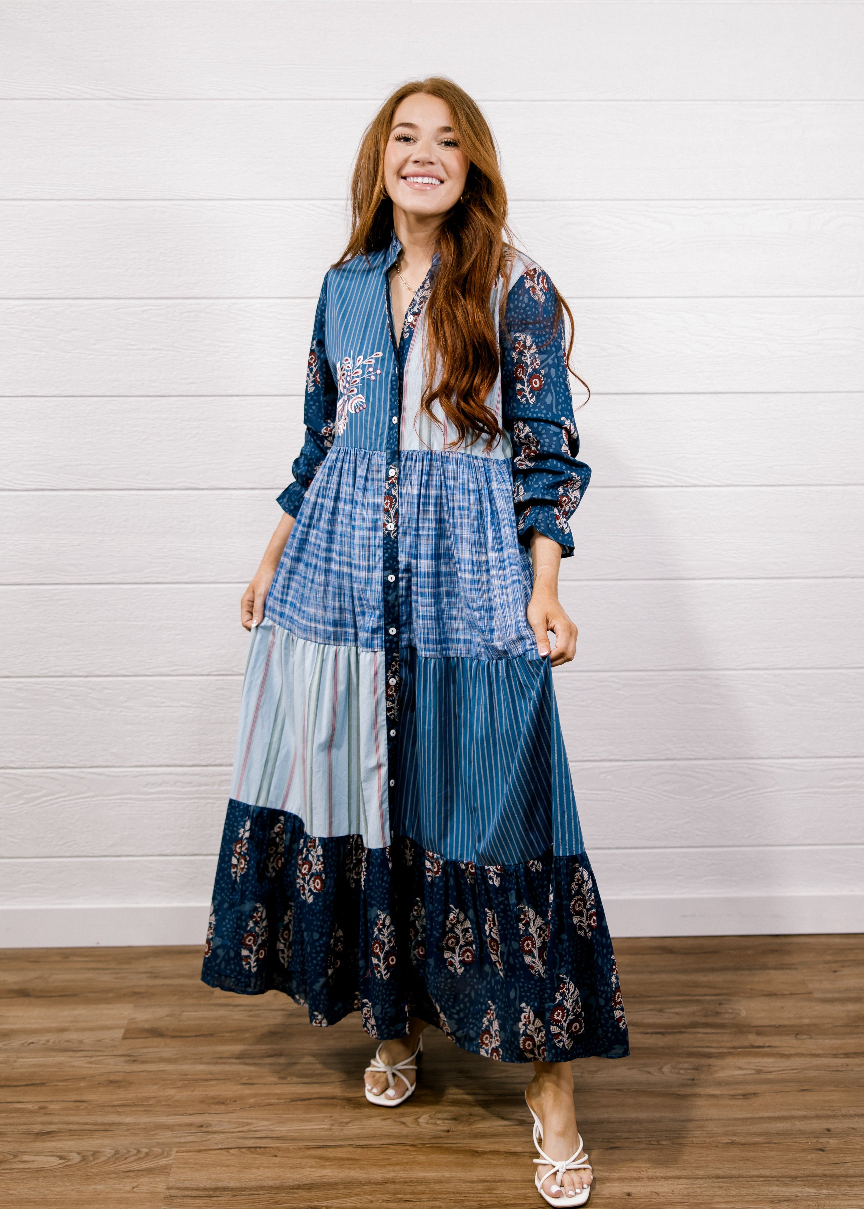 A woman modeling a long-sleeve patchwork maxi dress with blue plaid, striped, and floral prints, standing against a white shiplap wall on a wooden floor.