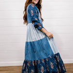 A woman modeling a long patchwork maxi dress with blue floral and striped panels, standing on a wooden floor against a white shiplap wall.