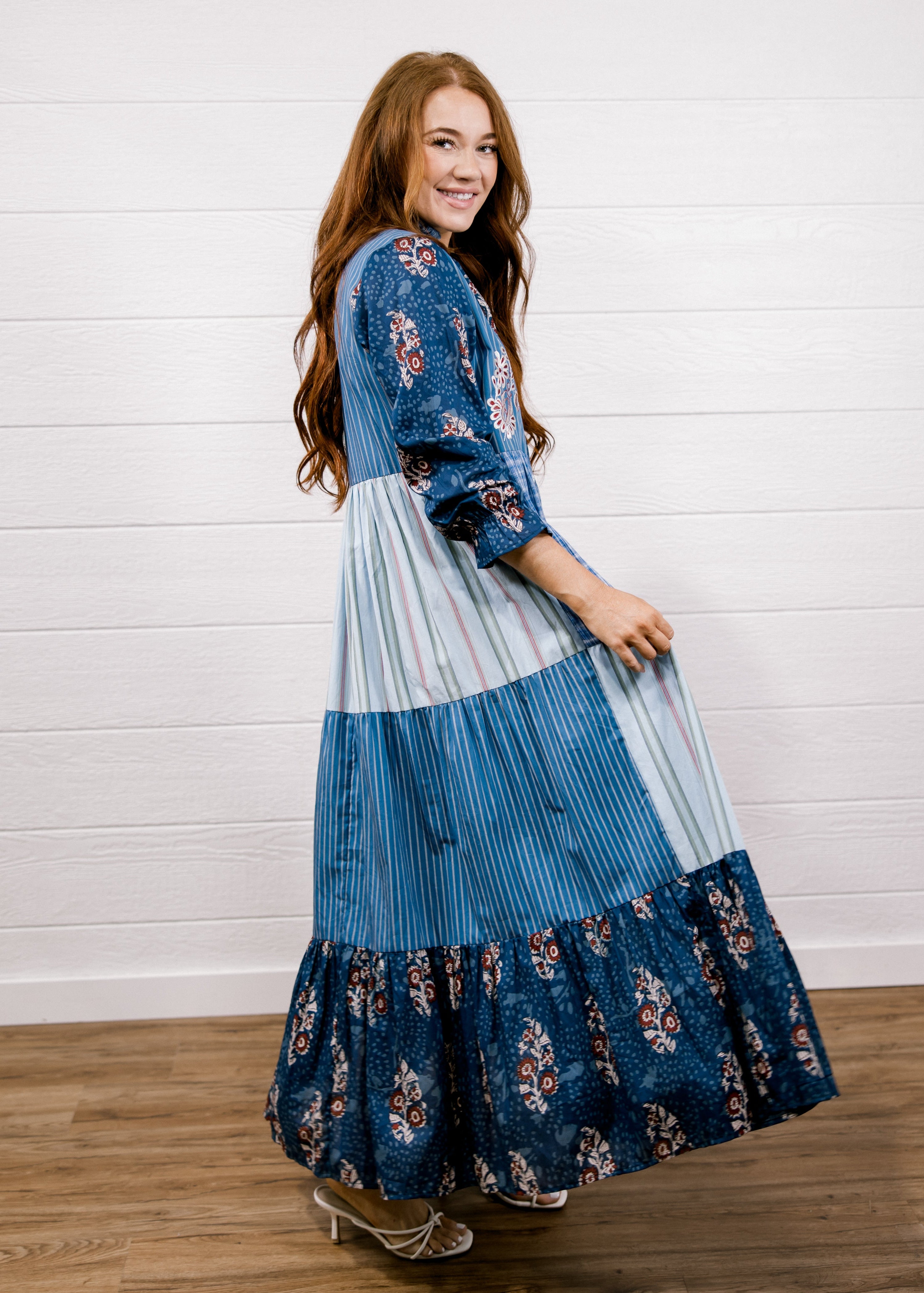A woman modeling a long patchwork maxi dress with blue floral and striped panels, standing on a wooden floor against a white shiplap wall.