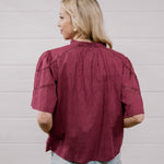 A woman with blonde hair is shown from the back wearing a short-sleeve, gathered blouse in a deep burgundy color, paired with light blue jeans, against a white paneled wall.