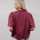 A woman with blonde hair is shown from the back wearing a short-sleeve, gathered blouse in a deep burgundy color, paired with light blue jeans, against a white paneled wall.