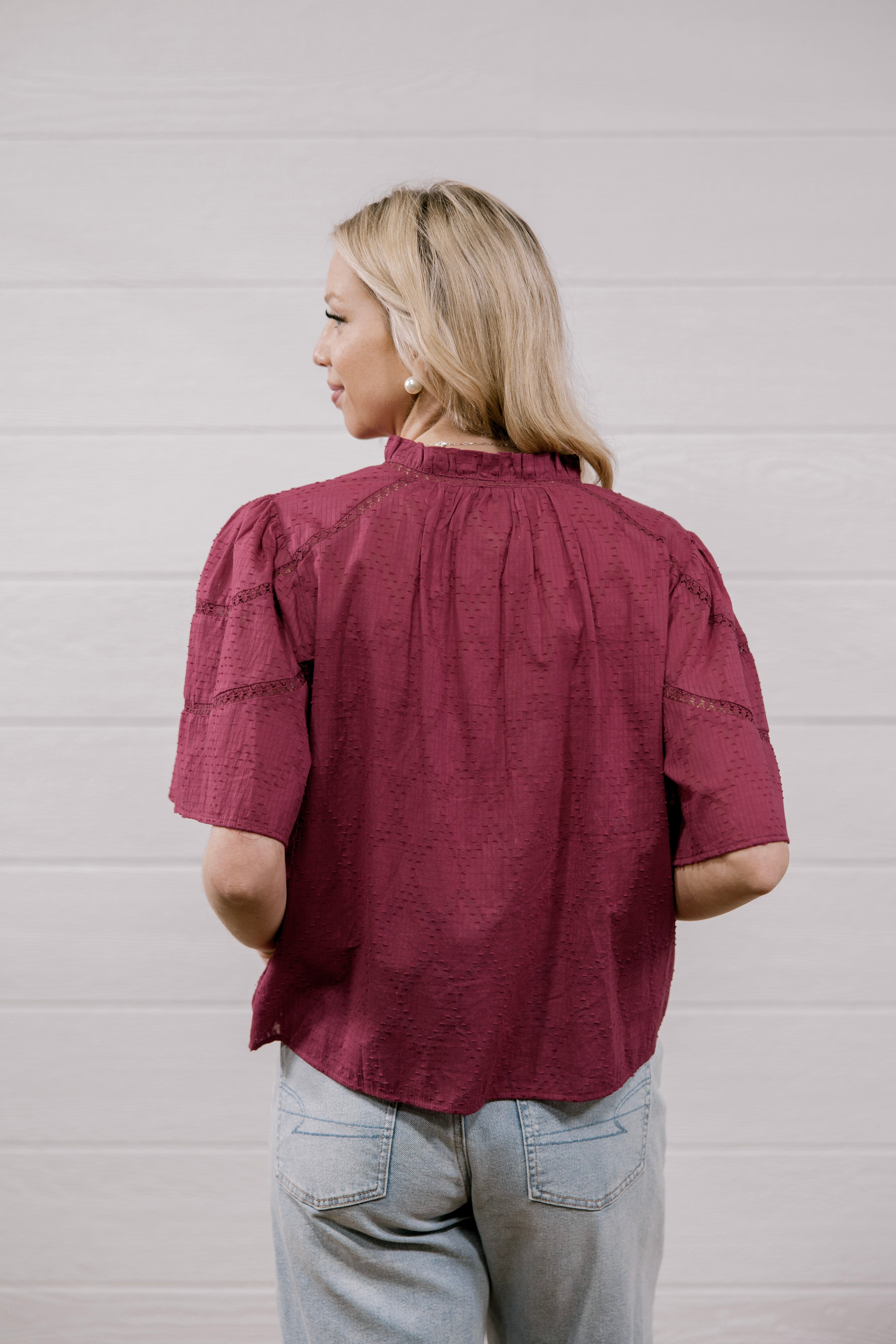 A woman with blonde hair is shown from the back wearing a short-sleeve, gathered blouse in a deep burgundy color, paired with light blue jeans, against a white paneled wall.