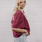 A woman with blonde hair wearing a burgundy short-sleeve button-up shirt and light wash jeans, standing against a white wood-paneled wall.