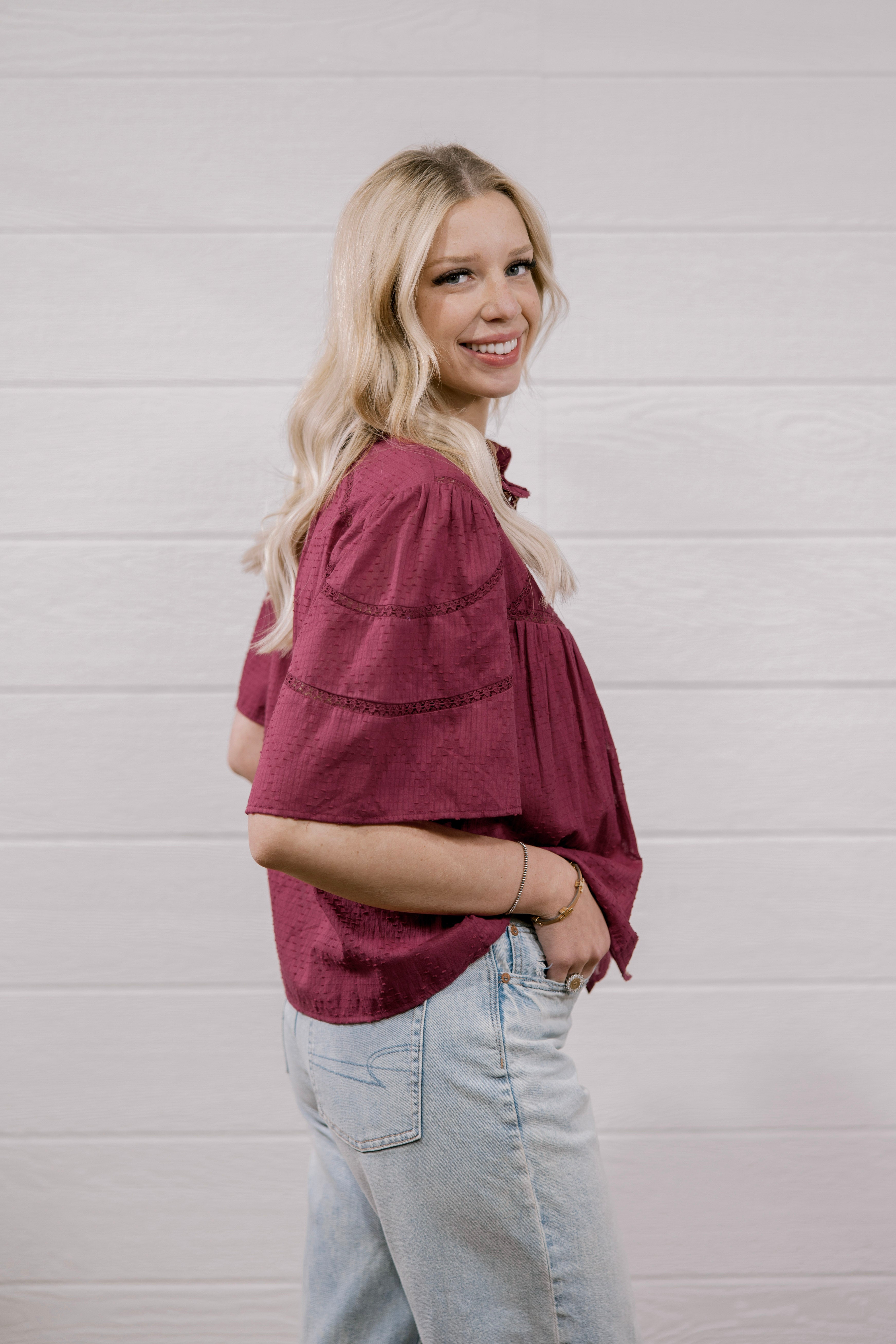 A woman with blonde hair wearing a burgundy short-sleeve button-up shirt and light wash jeans, standing against a white wood-paneled wall.