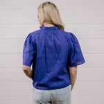 A woman viewed from behind wearing a purple textured short-sleeve blouse and light gray jeans, standing against a white paneled wall.