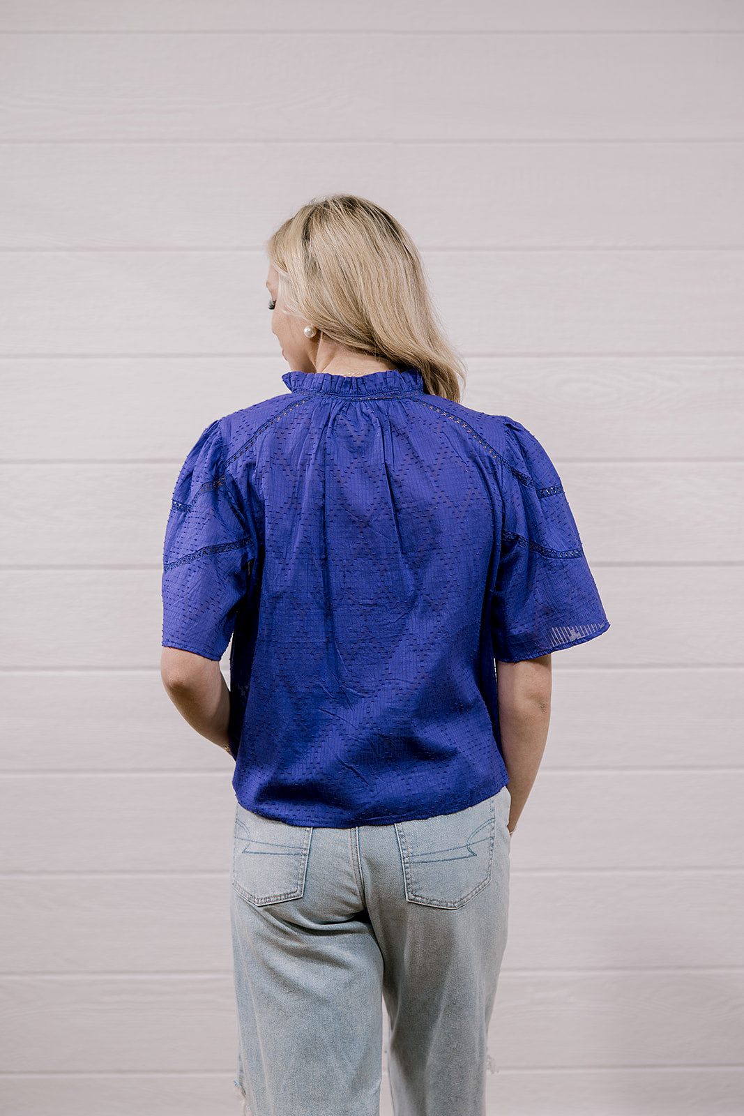 A woman viewed from behind wearing a purple textured short-sleeve blouse and light gray jeans, standing against a white paneled wall.
