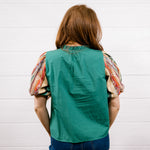 A woman viewed from behind wearing a green sleeveless vest over a short-sleeve blouse with patterned puff sleeves, paired with dark blue jeans, standing against a white wood-paneled wall.
