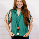 A smiling woman with long red hair stands against a white wood-paneled wall wearing a green short-sleeve blouse with patterned sleeves, dark blue jeans, and a long tassel necklace.