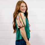 A woman with long red hair is shown from the side, wearing a green short-sleeve top with a colorful abstract print on the sleeves, paired with dark blue jeans, against a white wood-paneled wall.