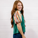 A woman with long red hair is shown from the side, wearing a green short-sleeve top with a colorful abstract print on the sleeves, paired with dark blue jeans, against a white wood-paneled wall.