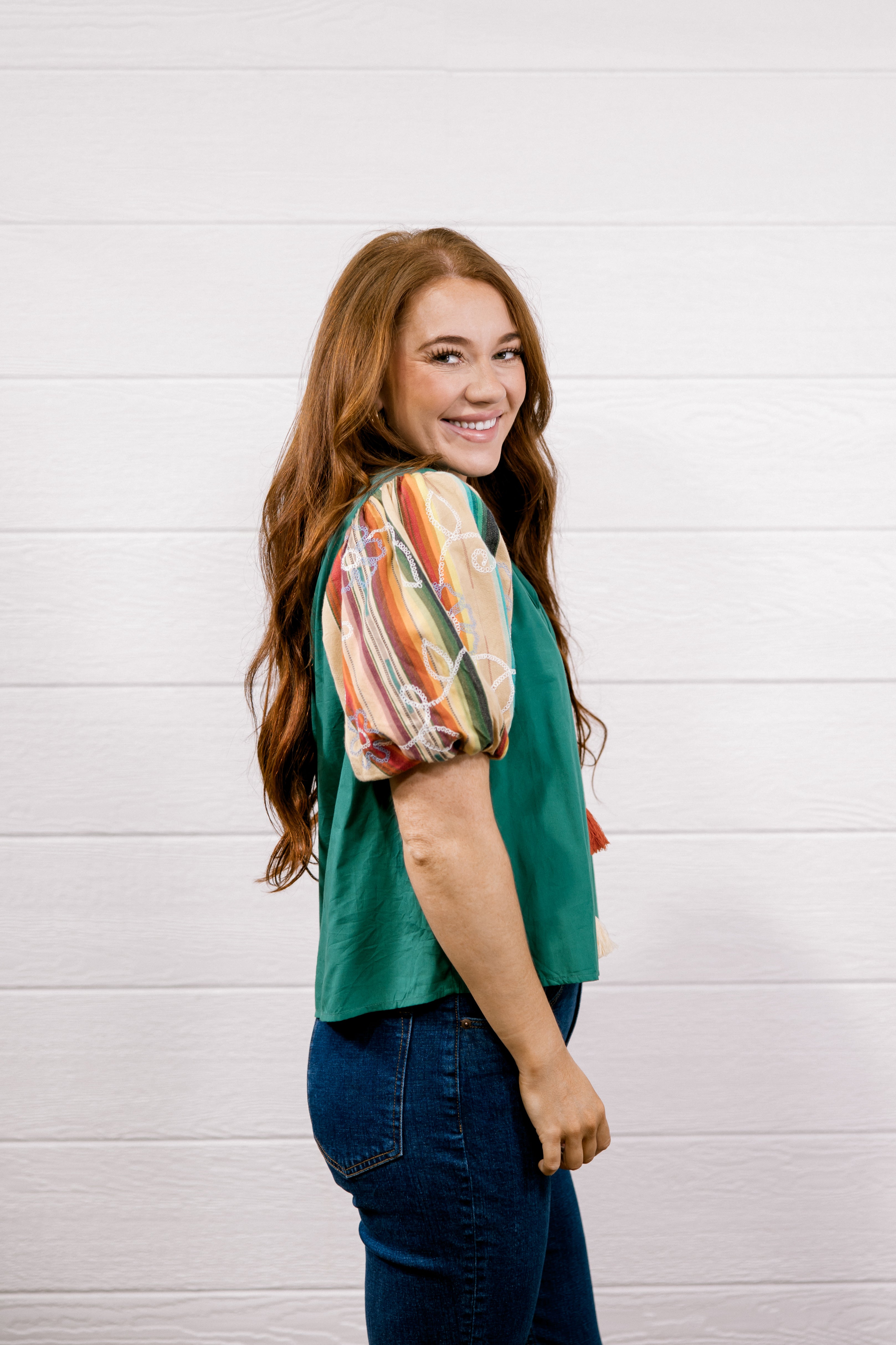 A woman with long red hair is shown from the side, wearing a green short-sleeve top with a colorful abstract print on the sleeves, paired with dark blue jeans, against a white wood-paneled wall.