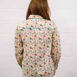 A woman viewed from behind wearing a long-sleeve western-style shirt with a beige background and a colorful cactus and floral pattern, paired with dark blue jeans, standing against a white wood-paneled wall.