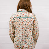 A woman viewed from behind wearing a long-sleeve western-style shirt with a beige background and a colorful cactus and floral pattern, paired with dark blue jeans, standing against a white wood-paneled wall.