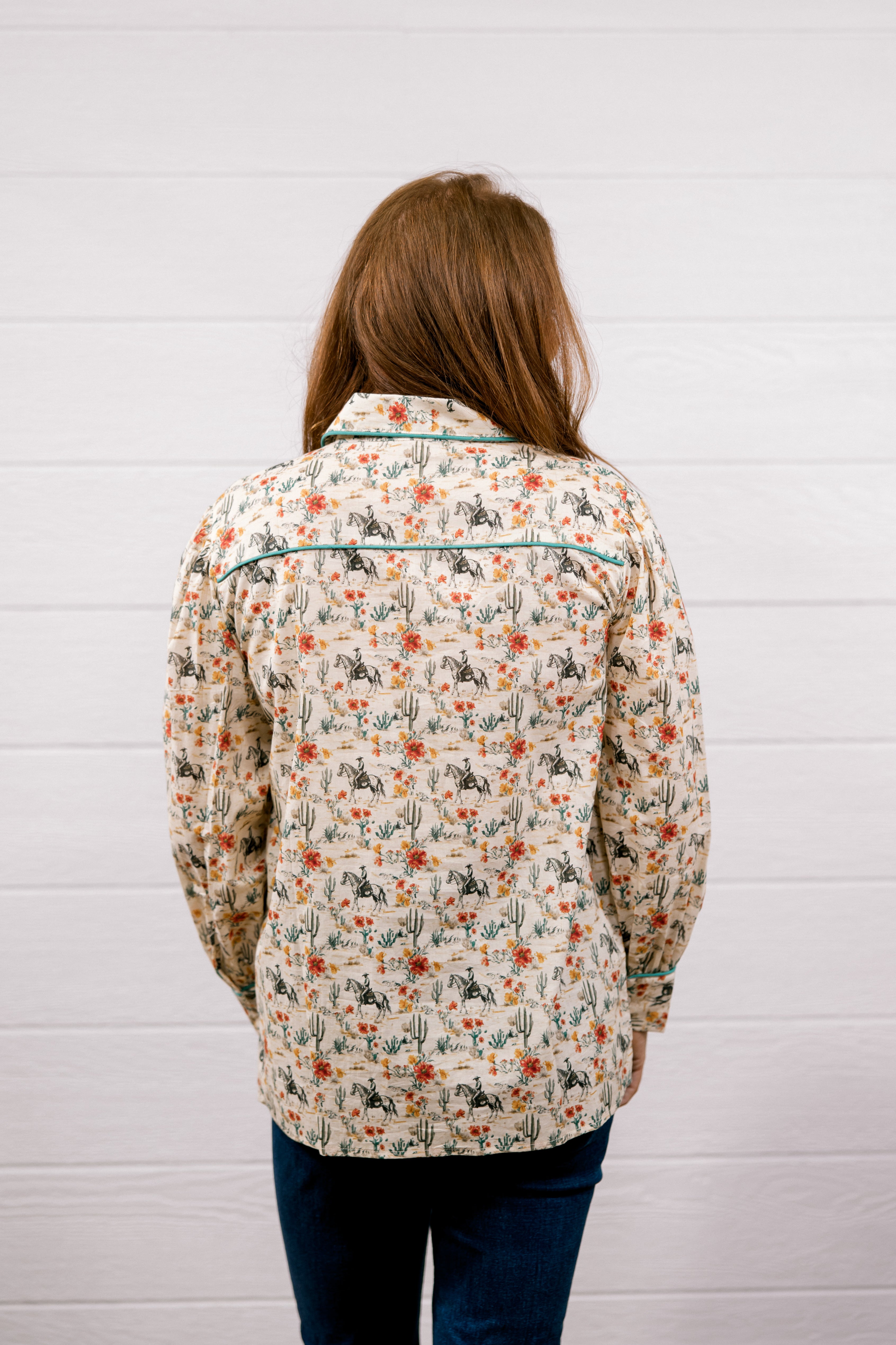 A woman viewed from behind wearing a long-sleeve western-style shirt with a beige background and a colorful cactus and floral pattern, paired with dark blue jeans, standing against a white wood-paneled wall.