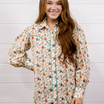 A smiling young woman with long brown hair stands against a white wood plank wall wearing a long-sleeve western-style button-up shirt with a beige base and a multicolor floral print, paired with dark blue jeans.