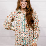 A smiling young woman with long brown hair stands against a white wood plank wall wearing a long-sleeve western-style button-up shirt with a beige base and a multicolor floral print, paired with dark blue jeans.