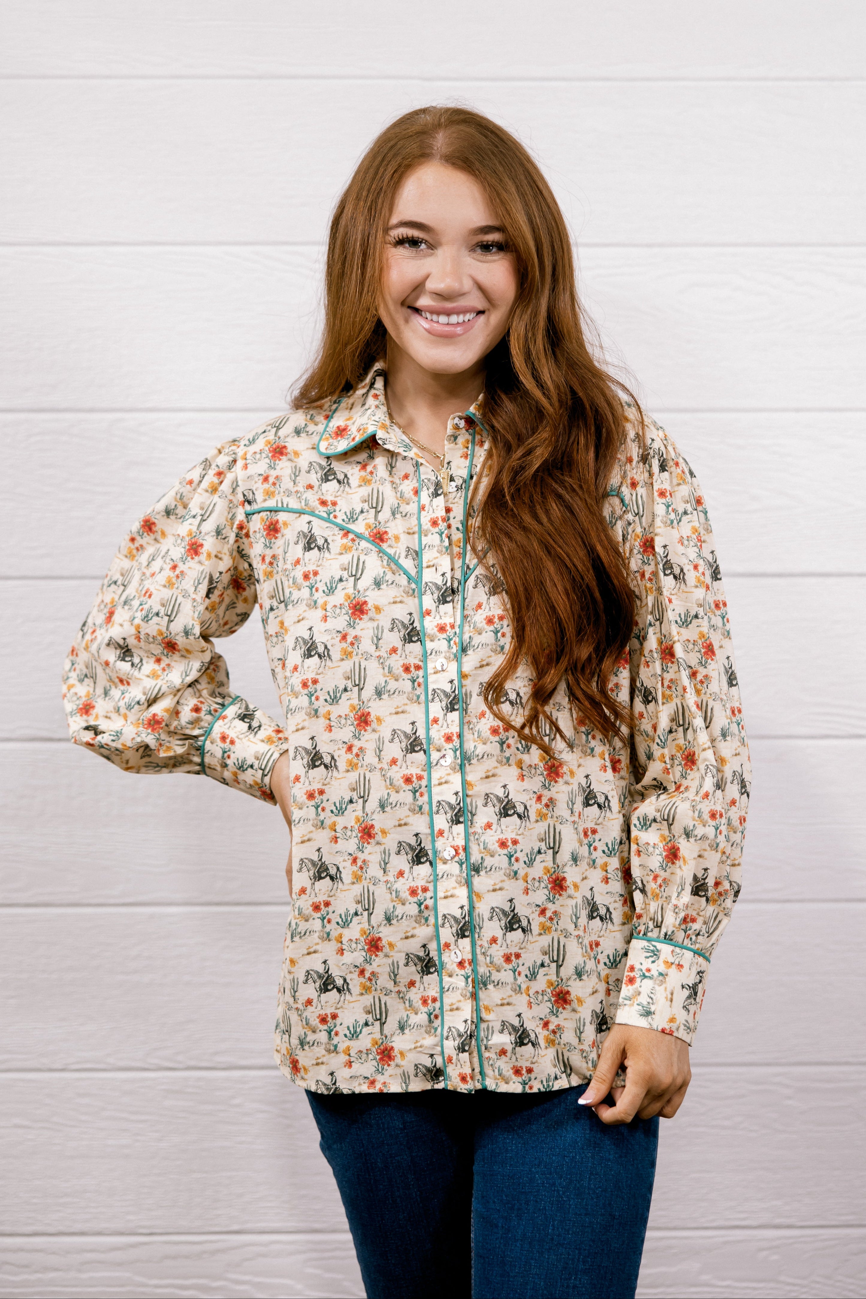 A smiling young woman with long brown hair stands against a white wood plank wall wearing a long-sleeve western-style button-up shirt with a beige base and a multicolor floral print, paired with dark blue jeans.