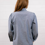 A woman viewed from behind wearing a blue and white vertically striped button-down shirt and dark blue jeans, standing against a white paneled wall.