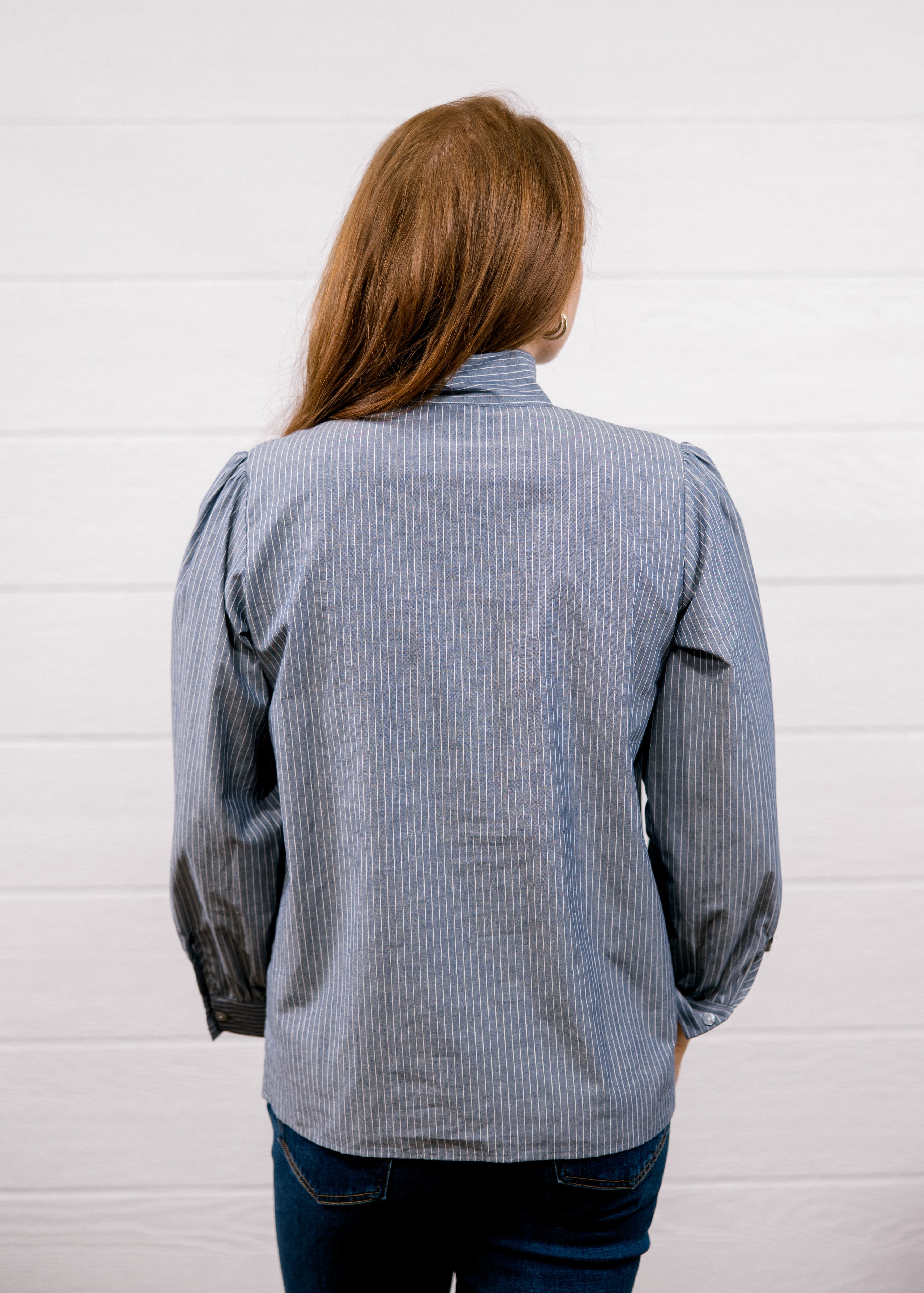 A woman viewed from behind wearing a blue and white vertically striped button-down shirt and dark blue jeans, standing against a white paneled wall.